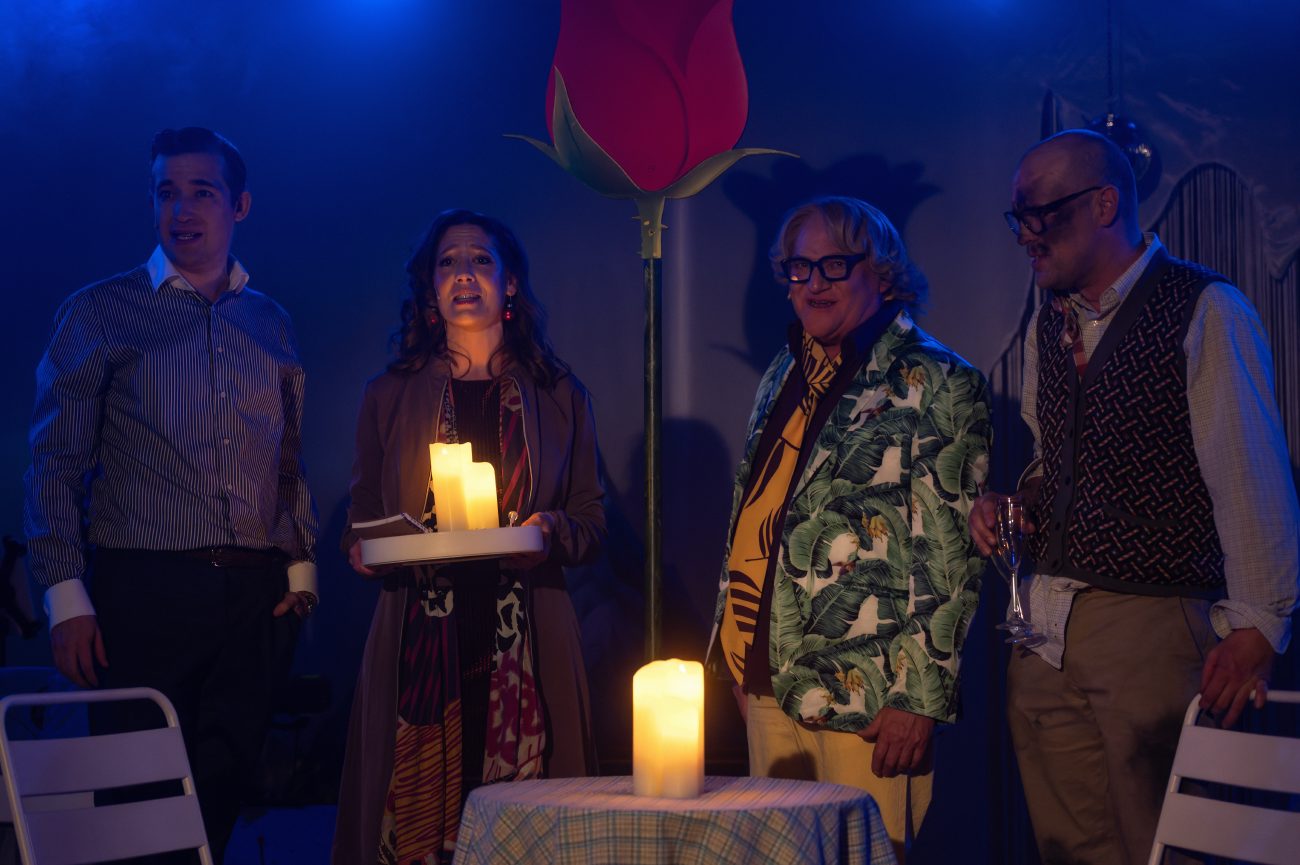 Mehrere Personen stehen dicht beieinander in einem Bühnenbild. Im Vordergrund sind ein Tisch mit Kerze und ein Glas zu sehen, im Hintergrund eine große stilisierte Rose. Einige Personen tragen Lichterketten, eine Person hält einen großen Strauß roter Blumen.
