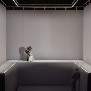 Eine große, helle Bühnenkonstruktion mit zwei Ebenen ist zu sehen. Oben links sitzt ein junger Mann, hinter ihm steht eine Frau; unten rechts steht ein Mann an der Wand. Die Bühne hat hohe graue Wände und leuchtende Linien an den Kanten.