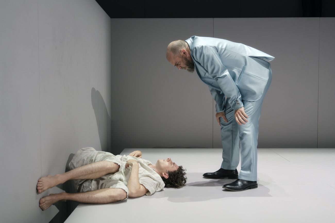Ein junger Mann liegt auf einer hellen Bühnenkante und blickt zur Seite. Er trägt helle Kleidung und liegt seitlich mit angewinkelten Beinen. Auf der Wand und der Bühnenkante sind grünliche und violette Projektionen zu sehen.