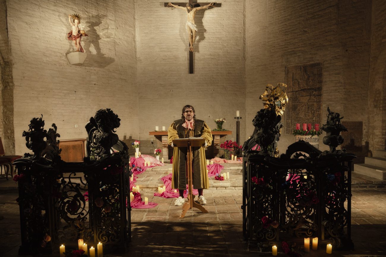 Eine einzelne Person steht in einer Kirche an einem Redepult vor einem Altar. Die Person trägt historische Kleidung, rundherum stehen Kerzen und pinke Stoffe auf dem Boden. Im Hintergrund sind Steinwände, ein Kreuz und religiöse Figuren zu sehen.