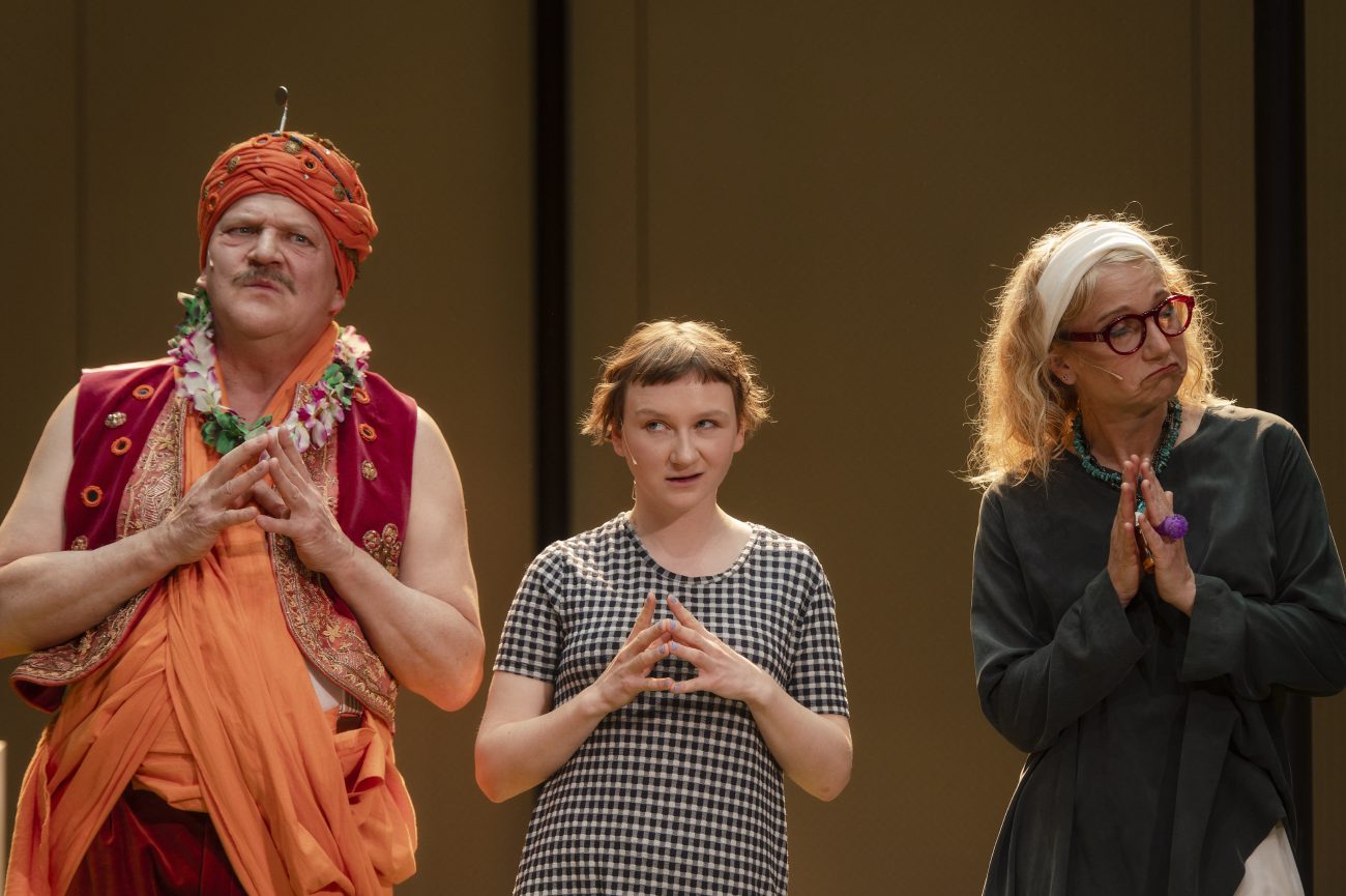 Ein Raum mit Bahnhofsuhren, Wänden und Rollrasen. Vorne rechts ein Mann in einem orangefarbenen indischen Kostüm, mit Schnurrbart und Turban, der beide Arme weit nach oben streckt, um ihn herum andere Personen, die ihn betrachten.